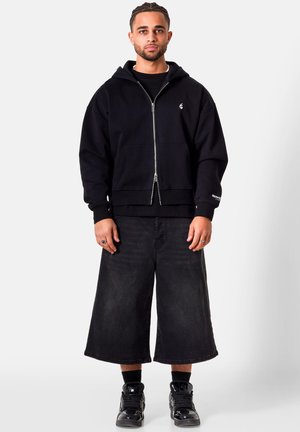 Mężczyzna stojący, ubrany w czarną bluzę z kapturem na suwak, szerokie czarne szorty do kolan, czarne skarpety i czarne trampki na jednolitym jasnym tle.