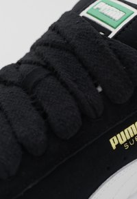 Gros plan sur des lacets noirs de baskets Puma et une partie de la languette avec le logo Puma vert et blanc, montrant le marquage doré Puma Suede sur le côté.