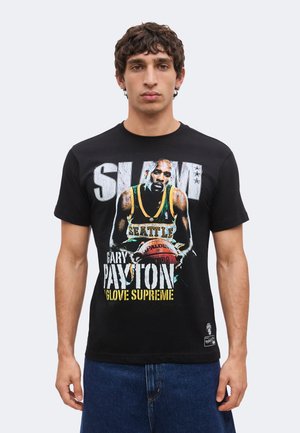 Mann trägt schwarzes T-Shirt mit Basketballspieler-Grafik und Text "SLAM Gary Payton Glove Supreme" und blaue Jeans vor schlichtem Hintergrund.