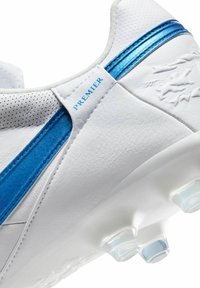 Botas de fútbol blancas con acentos azules, parte superior de cuero texturizado, detalles perforados y tacos transparentes en la suela para tracción.