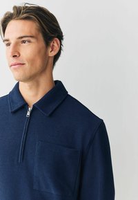 Giovane con capelli castano chiaro che indossa un top zip blu navy con coste, colletto e taschino sul petto, che guarda pensieroso verso sinistra.