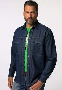 Camicia in denim blu scuro, con due tasche sul petto, chiusura con bottoni e maniche lunghe. Indossata sopra una camicia verde in tessuto testurizzato.
