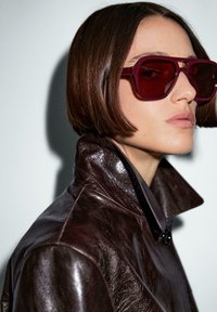 Braune Lederjacke mit hohem Kragen und einem Knopfdetail. Tiefrote, übergroße Sonnenbrille mit einer markanten rechteckigen Form.