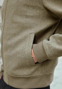 Beige strikjakke med tekstureret finish, ribmuffer og en sidelomme, der fremviser minimalistisk design med funktionelle detaljer.