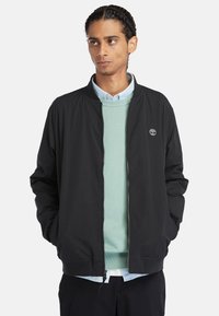 Giacca bomber nera con colletto alto, chiusura a zip e polsini elasticizzati, indossata sopra un maglione verde chiaro e una camicia blu con colletto.