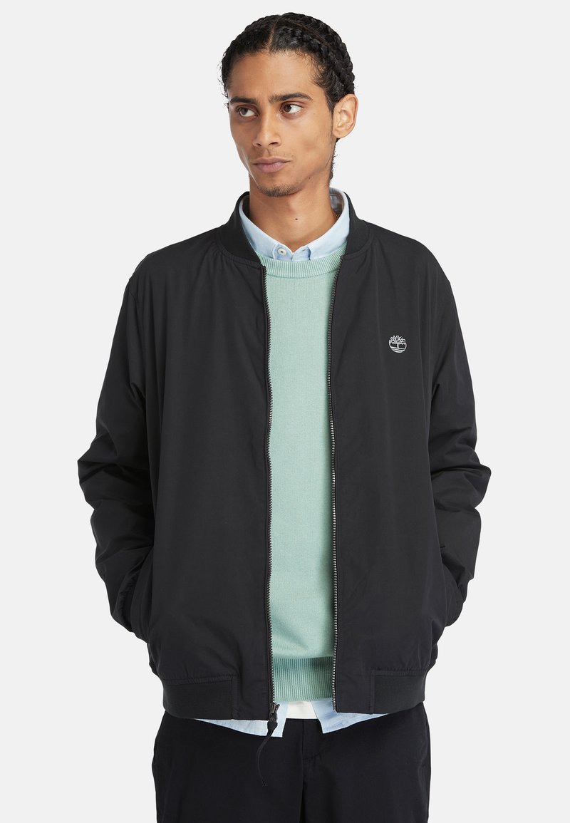 Giacca bomber nera con colletto alto, chiusura a zip e polsini elasticizzati, indossata sopra un maglione verde chiaro e una camicia blu con colletto.