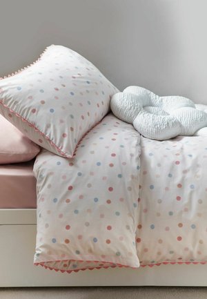 Bett mit weißer Bettwäsche mit mehrfarbigen Punkten, einem passenden Kissen und einem weißen, strukturierten Kissen in Knotenform vor einer einfarbigen hellen Wand.