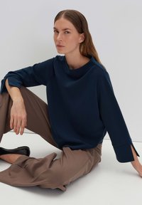 Navy blue long-sleeve top with a boat neckline and slits at the cuffs, paired with beige wide-leg trousers and black flats.