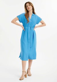 Abito azzurro chiaro, lungo fino al ginocchio, con maniche corte, scollo a V, dettaglio della cintura in vita e spacchi laterali. Indossato con sandali con tacco a blocco color cammello.