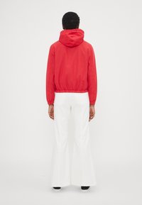 Czerwona kurtka z kapturem, z elastycznym pasem i mankietami, w połączeniu z białymi spodniami w stylu dzwonów. Tło jest jasne i jednolite.