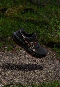 Črne športne čevlje z oranžnimi poudarki, teksturirano gumijasto podplato in oblazinjeno podporo za gleženj, zasnovane za tek po poteh ali zunanje aktivnosti.