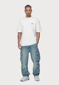 Biele oversized tričko s modrým grafickým potlačou, skombinované so svetlomodrými voľnými cargo džínsami s viacerými vreckami a uvoľneným strihom.