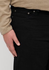 Sorte jeans med en glatt tekstur, med frontlommer og sølvfargede knappdetaljer. Hånden hviler på lårområdet.