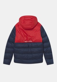 Padded jacket s červenou horní částí a tmavě modrou spodní částí. Obsahuje kapuci a diskrétní logo na zadní straně. Hladká, lesklá textura.