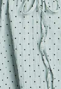 Svetlo modra tkanina z nagubano teksturo in črnimi pikami, ki ima zavezo na vratu in subtilne volančke ob robovih.