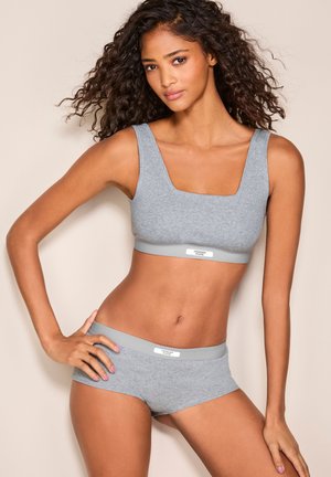 Ženska s kodrastimi lasmi, oblečena v ujemajočo se sivo rebrasto majčko brez rokavov in fantovske kratke hlačke, pozira z roko na boku pred bež ozadjem.