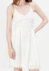 Une femme portant une robe en dentelle blanche sans manches avec de fines bretelles et une jupe évasée, se tenant avec une main sur la hanche devant un fond uni.