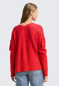 Rode oversized trui van een zachte, getextureerde stof met een V-achterkant en geribde zoom. Gecombineerd met losse blauwe jeans.