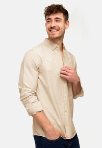 Hellbeige Knopfhemd, lange Ärmel, an den Manschetten hochgerollt, strukturierter Stoff, mit einer Brusttasche, kombiniert mit dunklen Jeans.