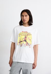 Hvid oversized bomulds T-shirt med et farverigt print af en springende figur sammen med "AIR JORDAN" og en bysilhuet i baggrunden.