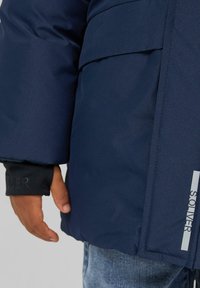 Marineblauwe pufferjack met een gestructureerde afwerking, voorzien van een klepzak en een zwarte manchet met branding. Zichtbare stiksels en een strak ontwerp.