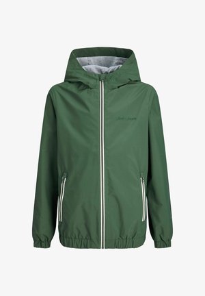 Lächelnder Junge mit gelber Mütze, grünem Reißverschluss-Jacke, weiter blauer Jeans und weißen Turnschuhen, stehend vor einem einfarbigen hellen Hintergrund.