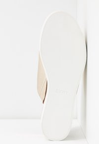Baskets beige clair en cuir texturé avec une semelle en caoutchouc blanche, arborant le logo "DKNY" au fond. Design et forme minimalistes.