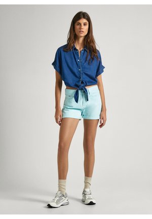 Denim marškiniai su mygtukais, sutrumpinti, su priekyje surišta juosta, derinami su mėtos žalios spalvos šortais. Modelis vilki pilkus sportinius batus ir kremine spalvos dryžuotas kojines.