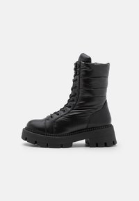 Botines negros de tobillo hechos de material sintético suave, con parte superior acolchada, cierre de cordones y una suela gruesa y texturada.