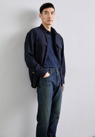 Navyblaues Hemd mit Knopfleiste und zwei Brusttaschen, kombiniert mit dunkelblauen Jeans. Einfaches Design, taillierte Passform, glatte Stofftextur.