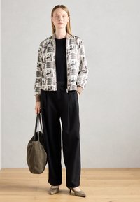 Giacca bomber con motivo beige, caratterizzata da stampe astratte, abbinata a una maglietta nera e pantaloni neri larghi. Tiene una morbida borsa verde oliva.