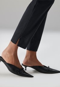 Zwarte leren mules met puntige neuzen en een decoratieve knoop. Slanke stilettohakken en openingen aan de uiteinden van zwarte taps toelopende broeken.