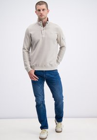 Heren lichtgrijze pullover met een gestructureerde afwerking, halve ritskraag en logo, gecombineerd met blauwe spijkerbroek en witte sneakers.