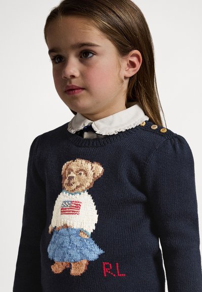 Temno modra pletena majica z grafiko medveda v belem puloverju z ameriško zastavo in denim krilo. Podrobnosti vključujejo rebrast ovratnik in gumbe.