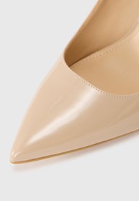 Pantofii cu toc înalt din piele verniculată bej au un vârf ascuțit și o finisare netedă, lucioasă. Interiorul este într-o nuanță mai deschisă, având un design închis.