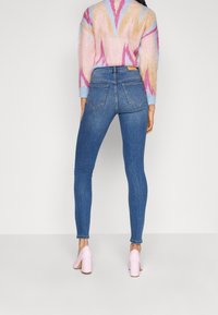 Vaqueros ajustados azules con textura de denim suave, cintura alta y dos bolsillos traseros, combinados con un suéter de punto multicolor y tacones rosa claro.