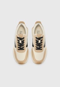Baskets de sport beiges et noires avec une tige synthétique lisse, un bout rond, des lacets noirs et des accents beiges contrastés sur la semelle intermédiaire.