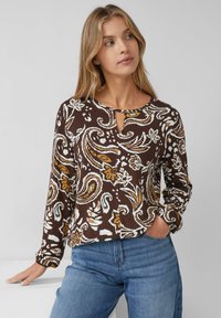 Kobieta z długimi blond włosami, ubrana w brązową, białą i musztardową bluzkę w wzór paisley oraz niebieskie dżinsy, pozująca z jedną ręką w kieszeni.