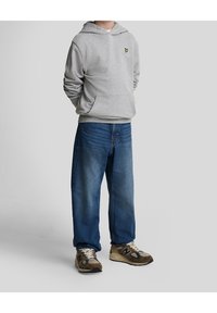 Szary hoodie z małym żółtym logo na klatce piersiowej, w zestawieniu z luźnymi niebieskimi dżinsami i brązowymi oraz beżowymi sneakersami.