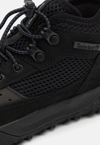 Zapato de senderismo negro con paneles de malla, acentos de ante suave y suela de goma duradera. Presenta cordones ajustables y un logotipo en el costado.