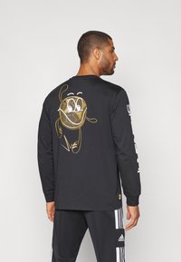 Camiseta negra de manga larga con gráfico de dibujos animados dorado en la parte posterior, con un diseño de malla. Texto blanco en la manga. Material de algodón suave.