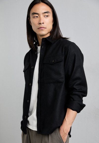 Black button-up shirt made of textured fabric, features two chest pockets and a slightly relaxed fit, paired with a white T-shirt underneath.