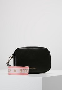Černá kožená crossbody taška obdélníkového tvaru, s zlatým logem a růžovým popruhem s bílým textem a akcenty.