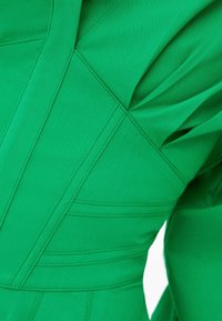 Zbliżenie na zielony materiał z geometrycznym przeszyciem i fałdami, ukazujące ustrukturyzowane szwy i szczegóły tekstury.