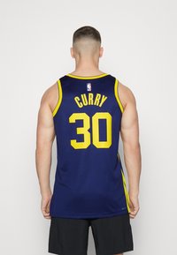 Koripallo-asu tummansinisellä taustalla ja keltaisilla yksityiskohdilla. Selkäpuolella lukee "CURRY" ja numero "30". Valmistettu kevyestä, hengittävästä kankaasta.