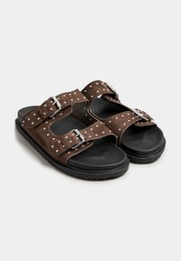 Sandali in suede marrone con doppie cinghie regolabili, borchie argentate e suola in gomma nera, caratterizzati da un design piatto e punta aperta.