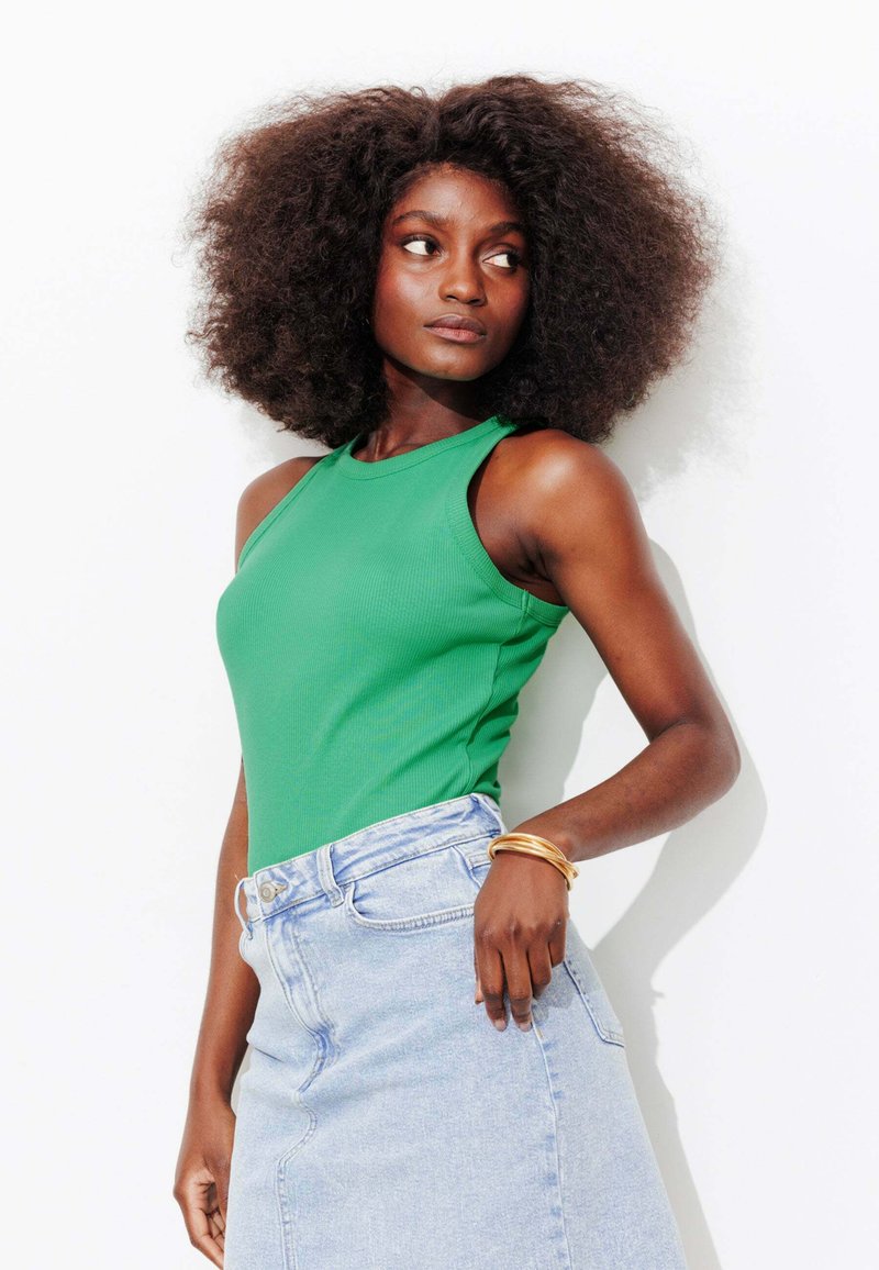 Femme aux cheveux bouclés et volumineux portant un haut vert sans manches et une jupe en jean bleu clair, posant devant un fond blanc.