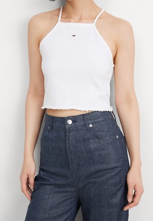 Person wearing a white halter crop top with a small logo and high-waisted dark blue jeans against a plain background.