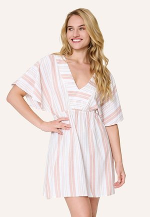 Žena s dlhými blond vlasmi sa usmieva, oblečená v krátkych, voľných šatách s výstrihom do V s ružovými a bielymi vertikálnymi pruhmi, ruka na bokoch.