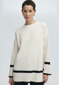 Bílý svetr s ribbed pleteninou, dlouhými rukávy, černými pruhy na lemech a manžetách a kulatým výstřihem, který má strukturovaný povrch.